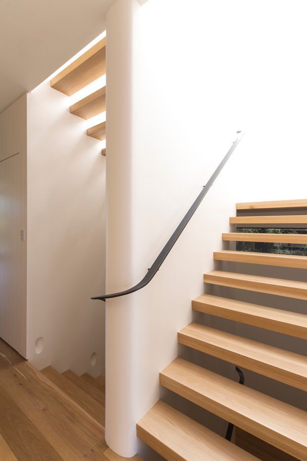 A bright, minimalist interior stairway with floating light timber treads and a slim dark metal handrail wrapping around a sculptural white cylindrical column. The stairwell includes open risers, a white plaster wall, warm timber flooring, and narrow timber ceiling beams or slats that allow light through. The overall composition is clean, refined, and contemporary.