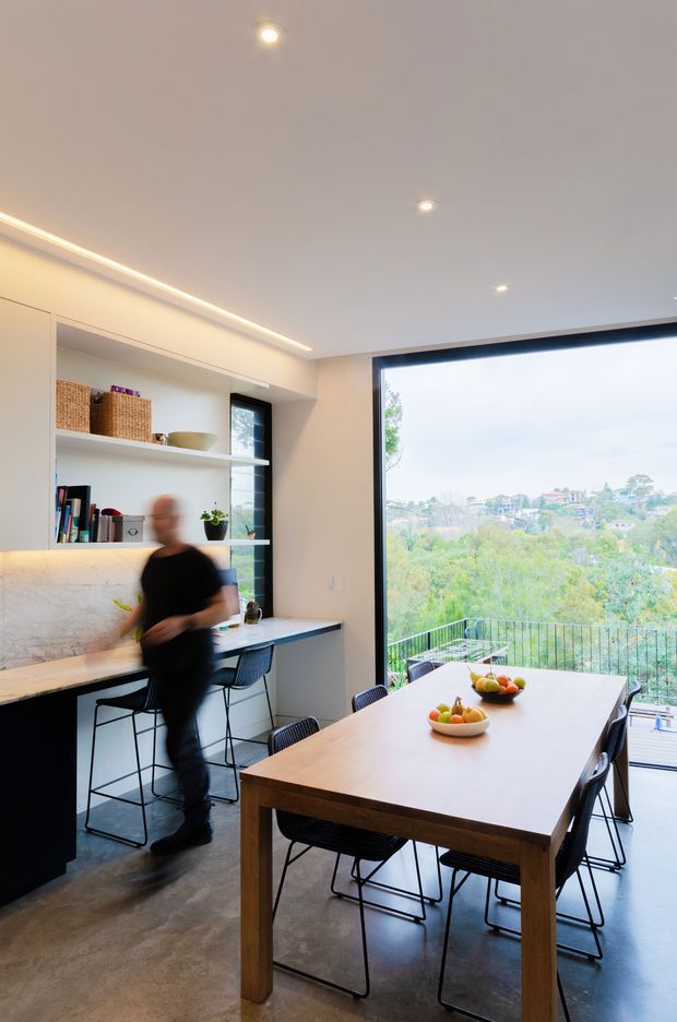 A bright, contemporary open-plan dining and kitchen space with a large timber dining table and black wire-frame chairs in the foreground. Along the left wall is a compact kitchen or work counter with dark lower cabinetry, a pale stone or marble-look backsplash, open white shelving, and integrated task lighting. A large floor-to-ceiling window and glazed door open to a balcony with a leafy suburban view. The space has a polished concrete floor, white walls and ceiling, recessed spotlights, and linear cove lighting.