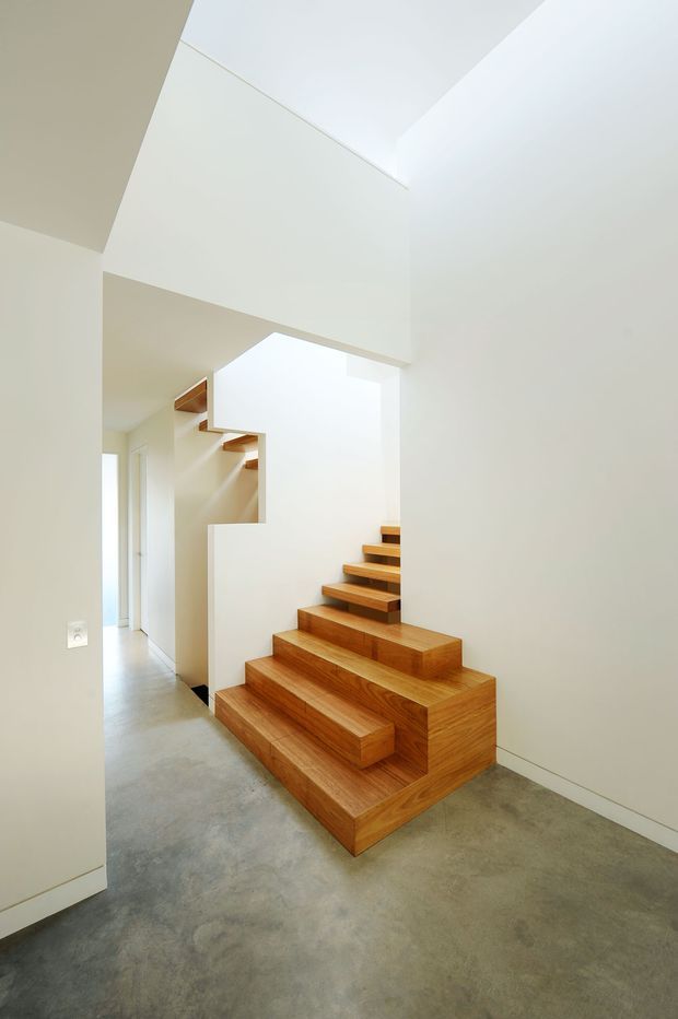 Bright minimalist interior view focused on a sculptural timber staircase. The stair has broad, box-like wooden treads and integrated stepped forms, set against smooth white walls and a high angular ceiling. A polished concrete floor extends through the hallway, with a small glimpse of adjoining circulation space and daylight.