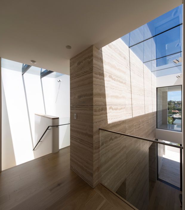 A refined contemporary stairway and landing with a sculptural central wall clad in large-format beige-brown stone or stone-look panels. Dark timber flooring, slim black metal handrails, white walls, recessed ceiling lights, and extensive floor-to-ceiling glazing create a bright, open architectural interior. Strong daylight enters through a glazed roof or clerestory, with exterior views visible beyond the stairwell.