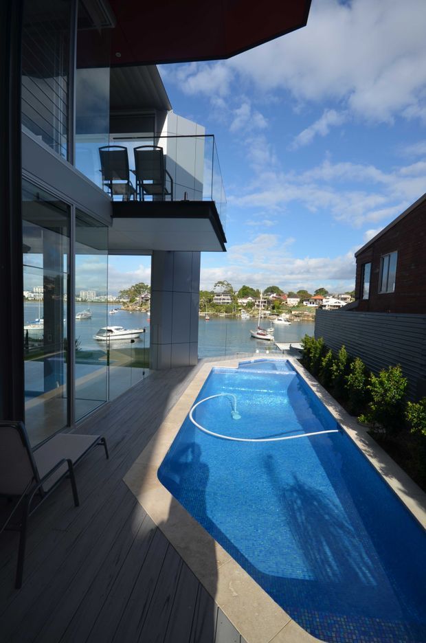 A narrow waterfront terrace with a long swimming pool beside a contemporary multi-level house. The pool is bordered by dark timber decking and pale paving, with full-height glazing, a cantilevered upper balcony, glass balustrades, and outdoor seating. A calm marina and houses are visible beyond the property. The scene has strong natural daylight, blue water, and modern architectural lines.