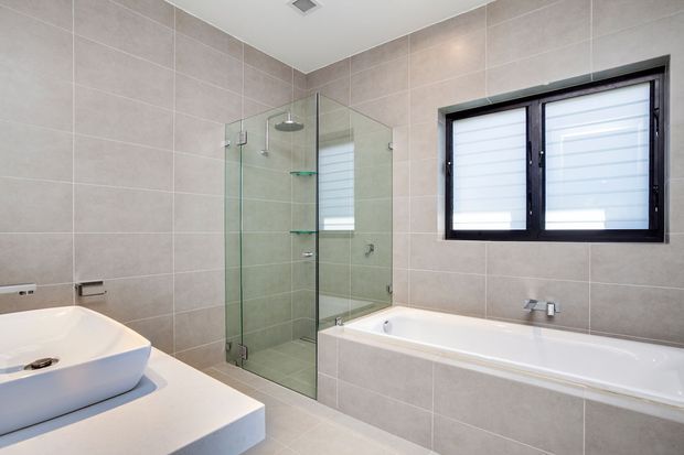 Contemporary bathroom with large light-grey tiled walls and floor, featuring a freestanding-style white bathtub, a glass-enclosed corner shower, a white vessel basin on a pale vanity, and a black-framed frosted louvre window. The space is clean, bright, and minimally styled.