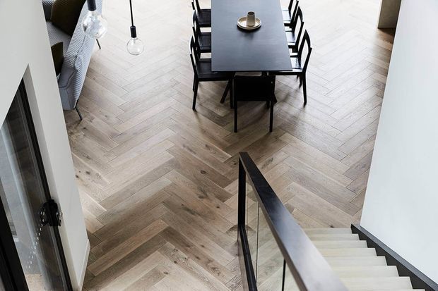 Elevated view over a refined dining area and stairway. A dark rectangular dining table with black chairs sits on a light timber herringbone floor, accompanied by simple pendant lights and a partially visible upholstered seating area. The foreground shows a modern staircase with pale treads, dark trim, and a slender black balustrade.