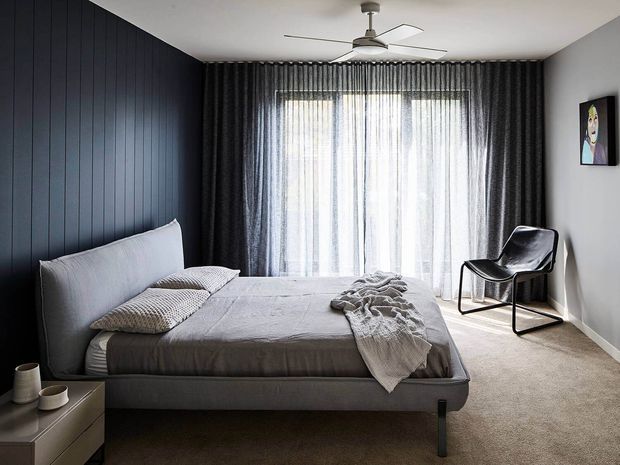 A contemporary bedroom with a large upholstered platform bed, dark vertical wall panelling, grey bedding, full-height sheer and dark curtains, carpeted flooring, a sculptural black lounge chair, and a ceiling fan. The room has a restrained monochromatic palette with strong natural light through the glazed opening.
