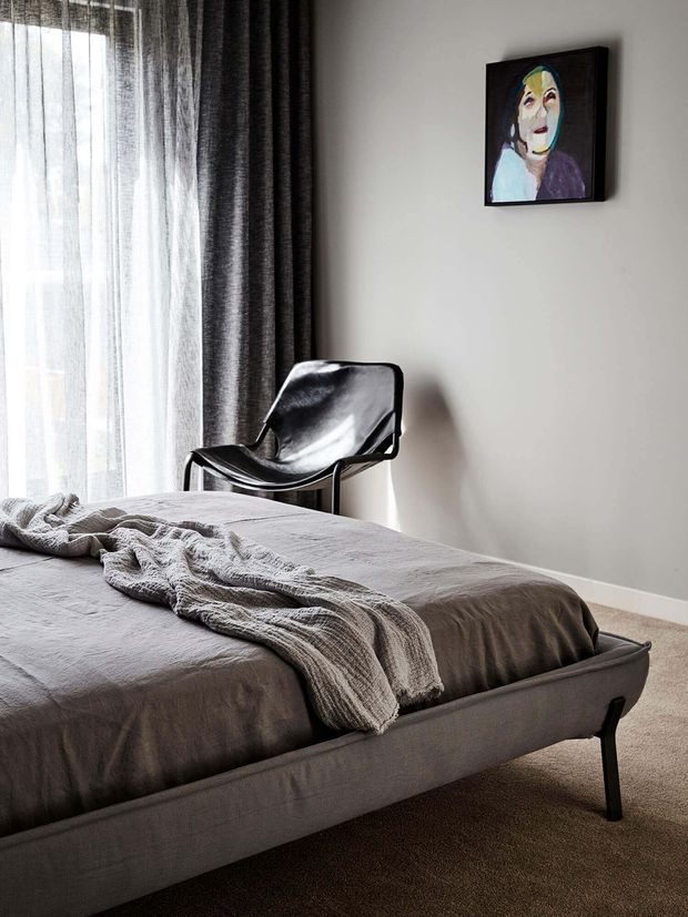 A calm, contemporary bedroom with a low upholstered bed dressed in grey bedding and a textured throw. A black leather sling chair sits beside full-height sheer and dark drapery, while a colourful framed portrait adds a small accent to the neutral room.