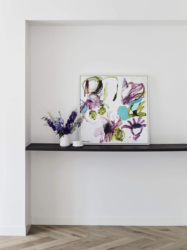 Minimal interior wall niche with a dark floating shelf displaying a framed abstract floral artwork and a small vase of purple flowers. The niche and surrounding walls are painted white, with light timber herringbone flooring and simple white skirting boards.
