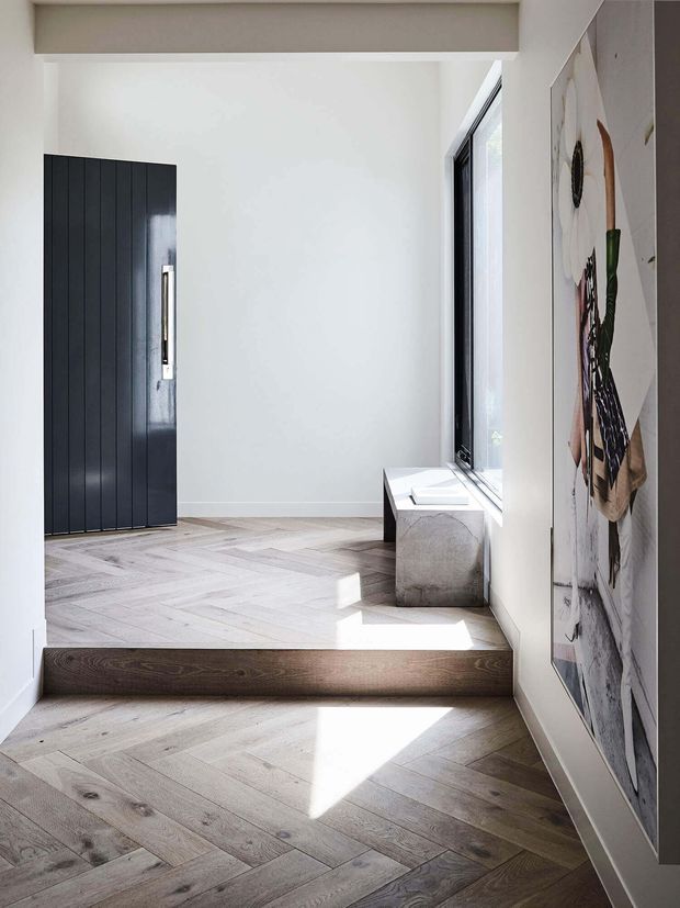 Bright minimalist residential entry hall with a single step leading to a raised landing. The space features pale herringbone timber flooring, white walls, a tall black vertically panelled entry door with a slim metal handle, a large black-framed window, a simple bench, and oversized wall artwork.