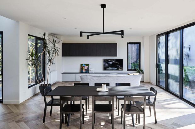 Bright open-plan dining and living area with a large dark timber dining table and matching chairs in the foreground. The room features pale herringbone timber flooring, white walls and ceiling, extensive black-framed glazing, a dark built-in wall cabinet, a low media console, contemporary artwork, indoor plants, and a sculptural black linear pendant light.