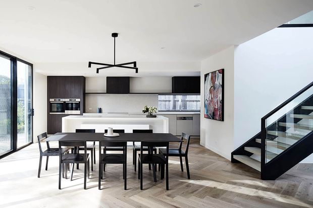 Bright open-plan dining and kitchen interior with a contemporary minimalist fit-out. A dark rectangular dining table with black chairs sits in the foreground, while the kitchen features dark timber cabinetry, a pale island bench, integrated appliances and a tiled splashback. Light herringbone timber flooring, white walls and ceiling, large glazed doors, a sculptural linear pendant, and a black-framed glass staircase complete the space. An abstract artwork provides a small colour accent.