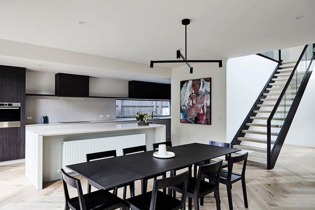 A contemporary open-plan kitchen and dining area with dark timber cabinetry, a white island bench, black dining table and chairs, and a striking black linear pendant light. A modern open staircase with black metal balustrade leads to the upper level, while an abstract artwork adds colour to the neutral interior.