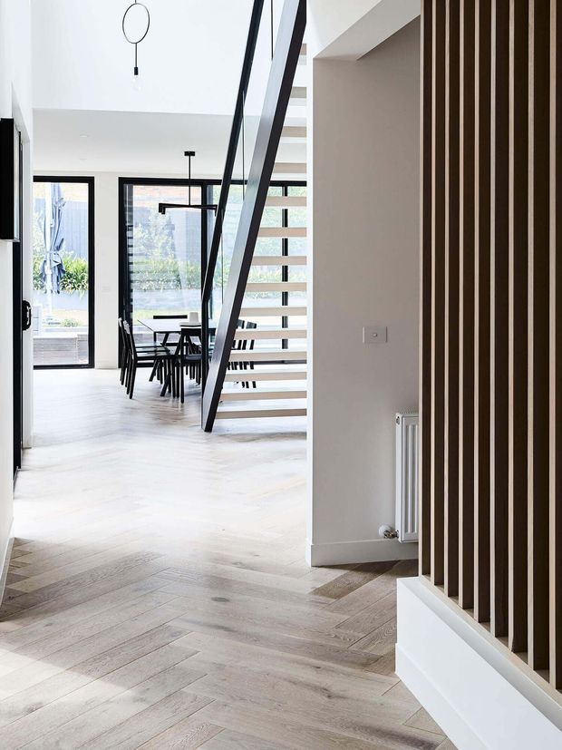 Bright contemporary residential interior viewed from an entry or hallway, with pale timber flooring, a floating open-riser staircase, black-framed glazing, a dining area beyond, and a prominent vertical timber slat feature screening built-in storage or the stair zone. The space has white walls, strong natural light, and minimalist black-and-white detailing.