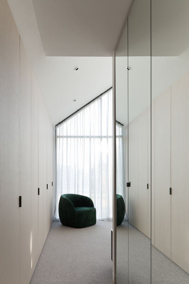 A narrow, softly lit dressing room or wardrobe corridor with full-height pale timber built-in cabinetry on both sides, a mirrored central door or panel, and a deep green upholstered lounge chair positioned in front of sheer curtains. The space has a high angled ceiling, minimalist recessed downlights, floor-to-ceiling glazing, and a light neutral carpet.