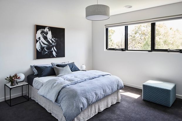 A bright, contemporary bedroom with a neatly made bed dressed in layered pale blue-grey bedding and dark accent cushions. The room has white walls, dark carpet, a large horizontal black-framed window, a grey drum pendant light, a small black side table, and a blue upholstered cube ottoman. A monochrome floral artwork provides a focal point.