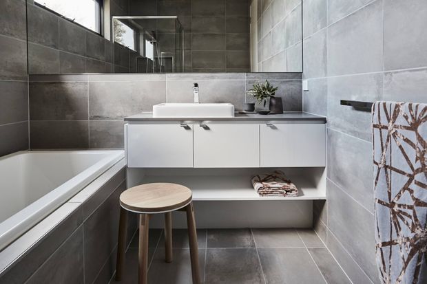 Contemporary bathroom with large-format grey wall and floor tiles, a white freestanding basin on floating white vanity cabinetry, bathtub, wall mirror, and minimal accessories. A wooden stool and patterned towel add warmth to the otherwise clean, neutral scheme.