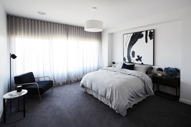 A refined contemporary bedroom with a large upholstered bed dressed in soft grey bedding, dark carpet, floor-to-ceiling sheer curtains, and a black bedside console. A dark lounge chair and small round side table sit by the window, while monochrome abstract artwork and sculptural lighting provide restrained decorative accents.