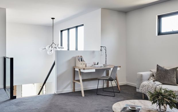 Bright contemporary upstairs workspace and sitting area overlooking a stairwell. The room has white walls and ceiling, dark grey carpet, black-framed windows, a minimalist timber desk with a task lamp and chair, a sculptural pendant light, and a light upholstered sofa with textured cushions. A small round coffee table and greenery complete the space.