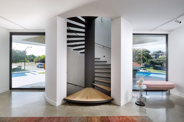 Contemporary open-plan entry hall featuring a sculptural curved staircase with floating timber treads and dark central structure. White curved walls frame the stairwell, while full-height glazing on both sides overlooks a swimming pool and landscaped suburban setting. The polished concrete floor and minimal furnishings create a clean, light-filled interior.