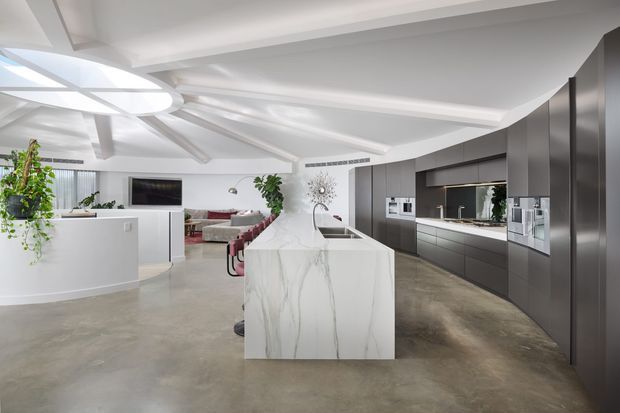Bright, open-plan contemporary kitchen with a large white marble-look island, dark charcoal full-height cabinetry, integrated appliances, and polished concrete flooring. The kitchen opens to a living area and features a sculptural white ceiling with recessed and angled forms, pendant-style integrated lighting, indoor plants, and a curved white reception-like divider.