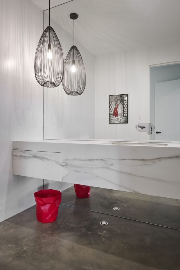 A contemporary bathroom viewed through a large wall mirror, featuring a floating double vanity clad in white marble-look material, a dark polished concrete floor with linear drains, and two sculptural red basin sinks. Two oversized black wire pendant lights hang above the vanity. White wall surfaces, a framed artwork, and a doorway are visible in the reflection.