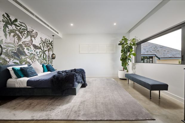 Bright contemporary bedroom with a low-profile dark upholstered bed, teal and navy bedding, a large botanical mural wall, pale carpet, a long upholstered bench, indoor planting, and wide horizontal glazing overlooking a neighbouring roof. The room has a clean white ceiling with recessed lighting and integrated air-conditioning.