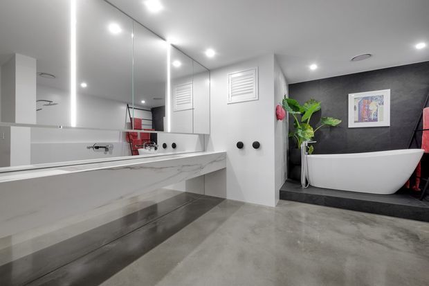Contemporary bathroom with a freestanding white bathtub set against a dark textured feature wall, large mirrored vanity cabinetry, a marble-look benchtop, polished concrete flooring, recessed ceiling lights, and indoor greenery.