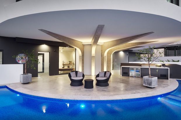 A luxurious modern indoor pool lounge with a curved pool edge in the foreground, pale stone flooring, sculptural arched structural columns, dark accent walls, integrated cabinetry, potted plants, and two contemporary lounge chairs. The space is brightly illuminated with concealed architectural lighting and features an open, airy layout.