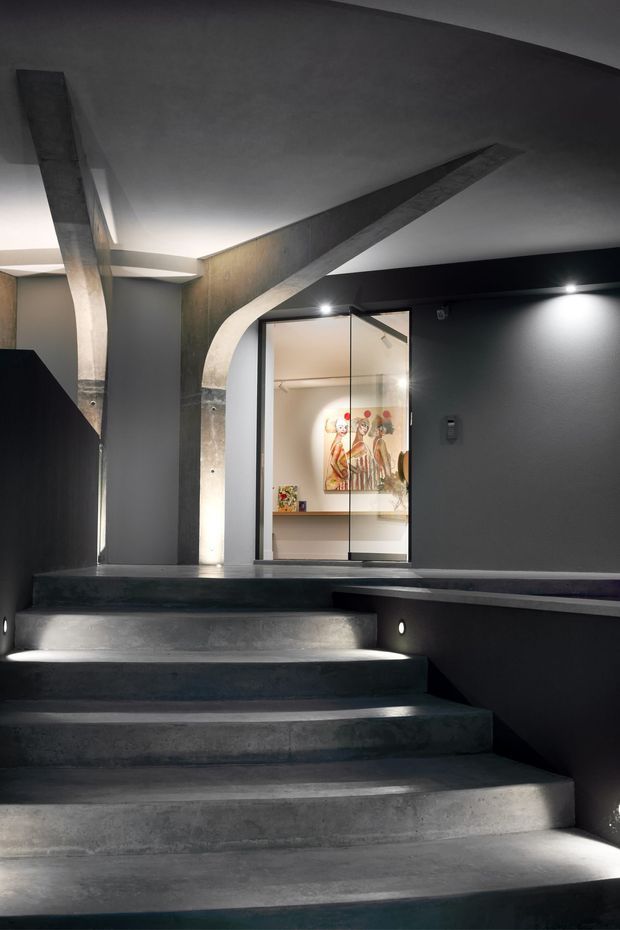 A dramatic contemporary stairway with broad poured-concrete steps, dark charcoal walls, integrated step lighting, and a sculptural curved concrete structural element rising toward the ceiling. A glazed doorway opens to a brightly lit interior space with artwork visible beyond.