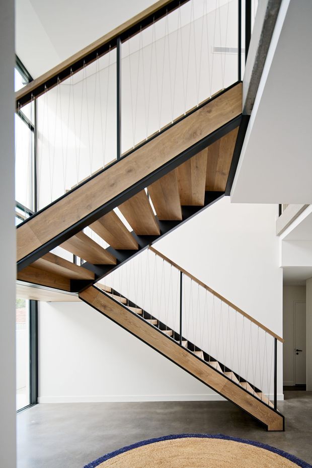 A bright contemporary interior featuring a sculptural multi-flight timber staircase with black structural framing and fine vertical cable balustrades. The stair rises through a double-height space with extensive floor-to-ceiling glazing, white walls, and a polished grey concrete floor. A partial woven rug is visible in the foreground.