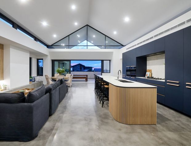 Open-plan contemporary kitchen and living area with a large curved island featuring vertical timber battens, dark navy cabinetry, integrated appliances, polished concrete flooring, and a high angular glazed ceiling feature. A dining area and living room sit beyond the kitchen, with large windows providing exterior views.