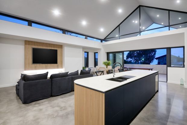 Open-plan contemporary kitchen and living area with a large dark island, white benchtop, integrated sink and tapware, timber slatted island end, polished concrete flooring, and a dark sectional sofa facing a timber media wall. Floor-to-ceiling glazing and a high glazed roofline bring in abundant natural light and views to the outdoors.