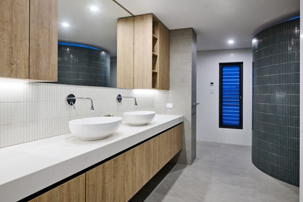 Contemporary bathroom with a long double vanity, white vessel basins, pale timber cabinetry, white vertical wall tiling, and a grey tiled floor. Curved dark tiled wall sections frame the space, while recessed lighting and a blue louvred window add contrast.