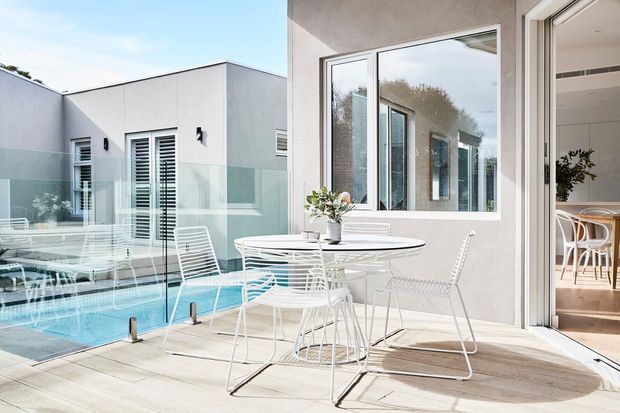 Bright contemporary outdoor terrace beside a swimming pool, furnished with a white metal dining table and chairs. Large glazed doors and windows connect the terrace to a light-filled interior, with a kitchen or dining area visible inside. The architecture features clean rectilinear forms, pale rendered walls, white shutters, and extensive glazing.