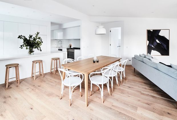 Bright open-plan interior combining a white kitchen, dining area, and living room. The space features pale timber flooring, white cabinetry and walls, a large light-wood dining table with white dining chairs, timber bar stools at the kitchen island, a pale sofa, pendant lighting, and abstract wall art.