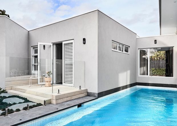 Contemporary white rendered residential exterior surrounding a bright blue swimming pool. The low-profile building features clean geometric volumes, white louvred doors and windows, dark base trim, a small paved terrace with outdoor seating, and a landscaped garden visible at the side.