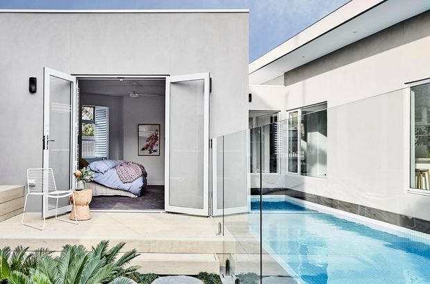 Modern white single-storey residence opening directly onto a narrow swimming pool courtyard. The image shows a bedroom through wide bi-fold doors, pale stone or concrete terrace steps, frameless glass pool fencing, expansive glazing, and a crisp flat-roofed architectural form.