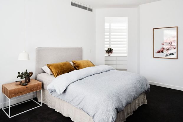 Bright, minimalist bedroom featuring a large upholstered bed with pale grey bedding, mustard accent pillows, a timber bedside table with white metal legs, white walls, plantation shutters, dark carpet, and simple framed artwork.