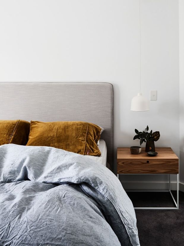 A softly styled contemporary bedroom featuring a large upholstered light-grey headboard, rumpled pale grey bedding, mustard-gold cushions, and a warm timber bedside table with minimalist metal legs. A simple white pendant light hangs beside the bed against a plain white wall, with a small plant and ceramic objects on the nightstand.