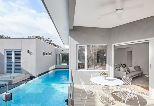 Bright contemporary outdoor entertaining area beside a narrow swimming pool, with a white-rendered flat-roof building, glass pool fencing, and a covered patio furnished with a round table and wire dining chairs. Large glazed doors open directly into a light living room.
