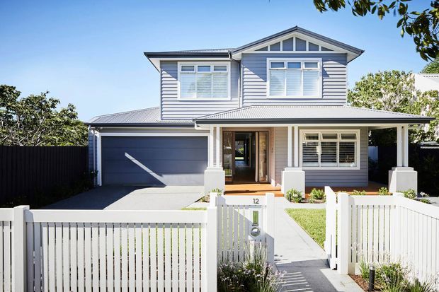 Bright daytime street-facing view of a two-storey suburban house with pale horizontal weatherboard cladding, a prominent gabled upper level, grey metal roofing, white-framed windows with shutters, an integrated single garage, and a white picket fence with a front garden and path.