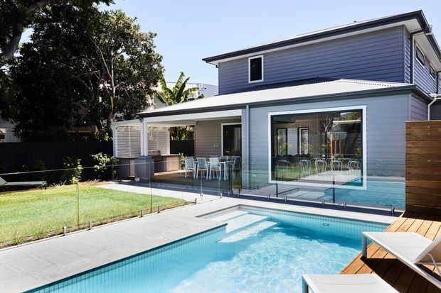 A contemporary two-storey residence viewed from the rear, with pale grey horizontal cladding, extensive glazing and a covered outdoor dining area opening onto a paved pool terrace. A rectangular swimming pool dominates the foreground, with timber decking and lounge chairs at the side, while a private lawn and mature planting border the property.