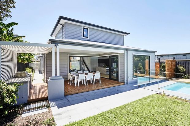 A contemporary two-storey residential home with a light grey weatherboard facade, covered outdoor dining area, timber decking, landscaped courtyard, and a clear-glass pool fence enclosing a rectangular swimming pool. The rear facade features large glazed openings connecting the interior living space to the outdoor entertaining area, while a pergola and slatted screening provide shade and privacy.