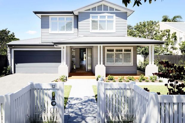 Front exterior view of a two-storey suburban home with a symmetrical gabled form, grey horizontal weatherboard cladding, white trim, a broad covered front porch, attached double garage, and white picket fencing. The landscaped front path leads to a recessed entry.