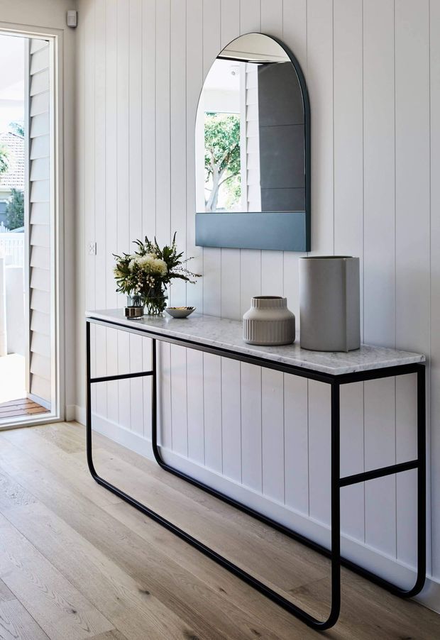 A bright, refined entryway featuring white vertical wall panelling, pale timber flooring, and a slim black metal console table with a light stone-look top. An arched black-framed mirror hangs above the console, which is styled with a vase, tall cylindrical vessel, and floral arrangement. A glazed exterior door with louvred detailing is visible to the left.
