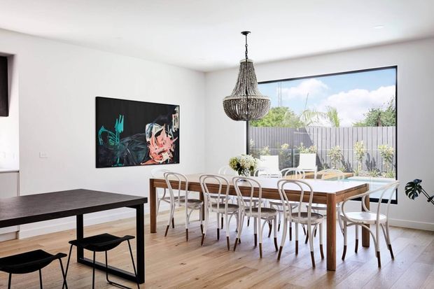 Bright contemporary open-plan dining area with a long timber dining table surrounded by curved-back chairs, a dark rectangular table in the foreground, light timber flooring, white walls, a large garden-facing window, and a prominent woven pendant chandelier. Abstract wall art and indoor plants add visual accents.