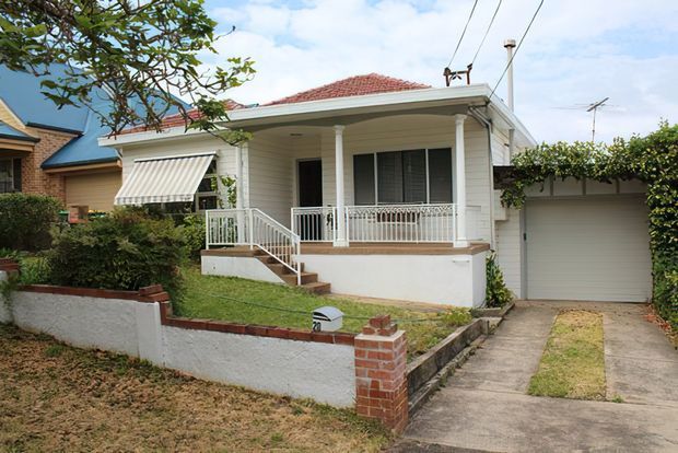 Street-facing view of a traditional single-storey suburban house with a white weatherboard facade, low-pitched red tiled hipped roof, covered front verandah supported by slender columns, decorative white balustrade, front steps, awning-covered window, and an attached single garage. A small front garden, low masonry boundary wall, and concrete driveway are visible.