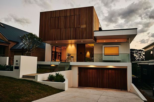 Front exterior view of a contemporary multi-level residence with a large vertically clad timber facade, crisp white rendered walls, broad flat roof forms, recessed warm lighting, upper-level glazing and a timber garage door. The home sits on a sloping suburban site with a concrete driveway and landscaped front garden.