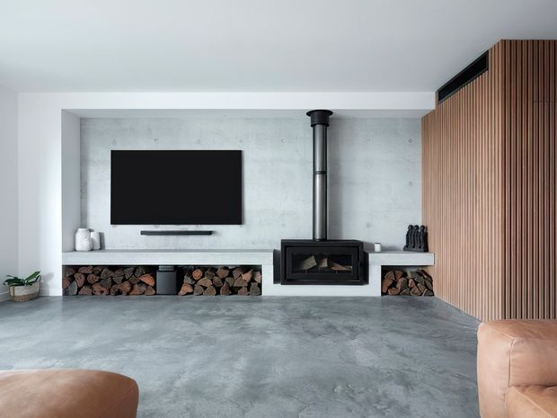 Modern minimalist living room centred on a built-in concrete fireplace wall with a black wood-burning stove, exposed flue, recessed firewood storage, and wall-mounted television. The room has a polished grey concrete floor and matching concrete wall surfaces, with a vertical timber slat feature wall on the right and partial tan leather seating in the foreground.