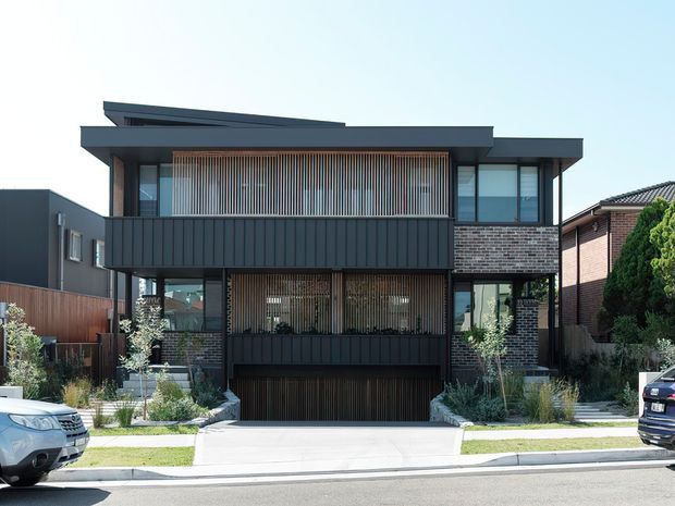 Contemporary two-storey urban residence viewed from the street, featuring a dark standing-seam style facade, extensive glazing, exposed brickwork, vertical timber screening, and a layered low-pitched roofline. The house includes an integrated lower-level garage, landscaped front garden, paved driveway, and neighbouring suburban homes.