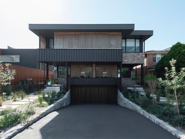Front elevation of a contemporary multi-storey residence with a broad flat roof, dark vertical cladding, exposed brickwork, timber screening, and extensive glazing. A central driveway leads to an integrated double garage, with landscaped garden beds and low stone retaining walls framing the entry.