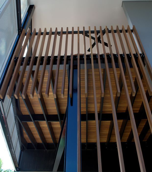 Upward-looking view of a contemporary architectural interior featuring a dramatic double-height void, exposed black structural framing, warm timber soffit panels, and a vertical timber slat screen. Tall glazing with horizontal blinds lines one side, while a dark blue structural column rises through the space.